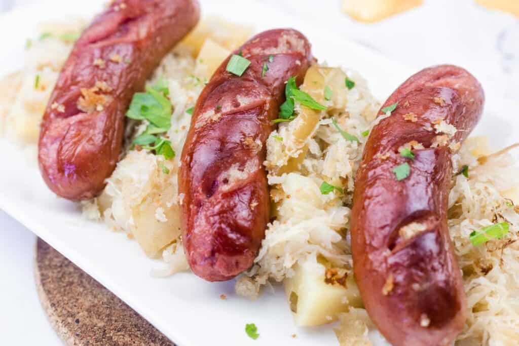 Three grilled sausages served on a bed of sauerkraut and diced potatoes, garnished with chopped parsley on a white rectangular plate.