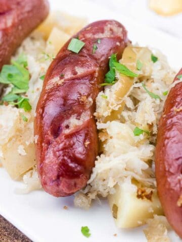 Three grilled sausages served on a bed of sauerkraut and diced potatoes, garnished with chopped parsley on a white rectangular plate.