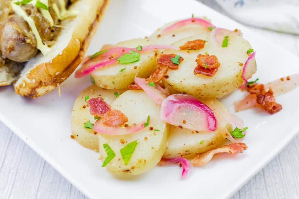 Sliced potatoes with bacon, onions, and herbs served on a white plate next to a sausage in a bun.