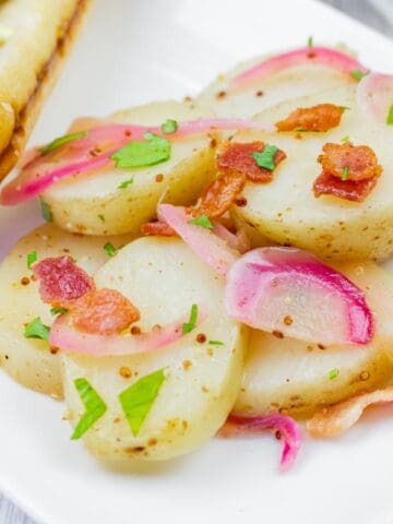 Sliced potatoes with bacon, onions, and herbs served on a white plate next to a sausage in a bun.