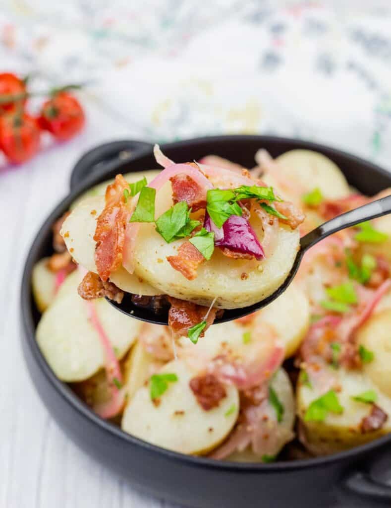A black spoon holds sliced potatoes, bacon, onions, and parsley above a bowl filled with the same potato salad.