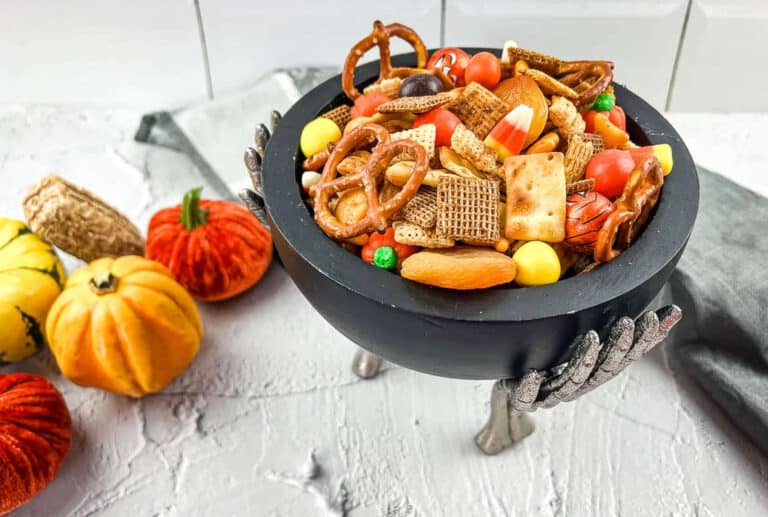 A black bowl held by silver skeleton hands filled with snack mix, pretzels, candy corn, and crackers, with small decorative pumpkins nearby.