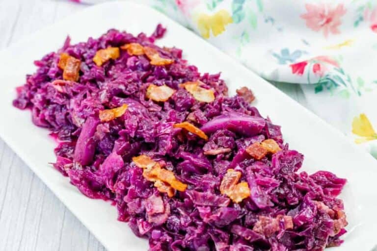 A rectangular white plate with cooked red cabbage mixed with bacon pieces, set on a light wooden surface with a floral napkin nearby.