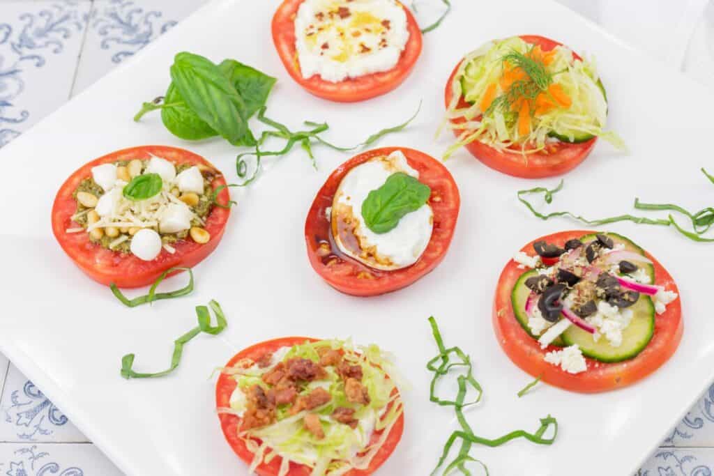 Sliced tomatoes topped with various cheeses, vegetables, and herbs, arranged on a white plate with basil and shredded greens as garnish.