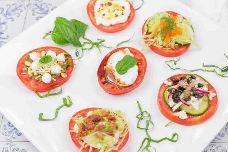 Sliced tomatoes topped with various cheeses, vegetables, and herbs, arranged on a white plate with basil and shredded greens as garnish.