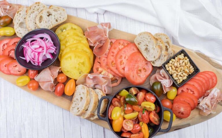 A wooden board with sliced bread, yellow and red tomatoes, assorted cherry tomatoes, prosciutto, pickled onions, pine nuts, and a small bowl of tomatoes.