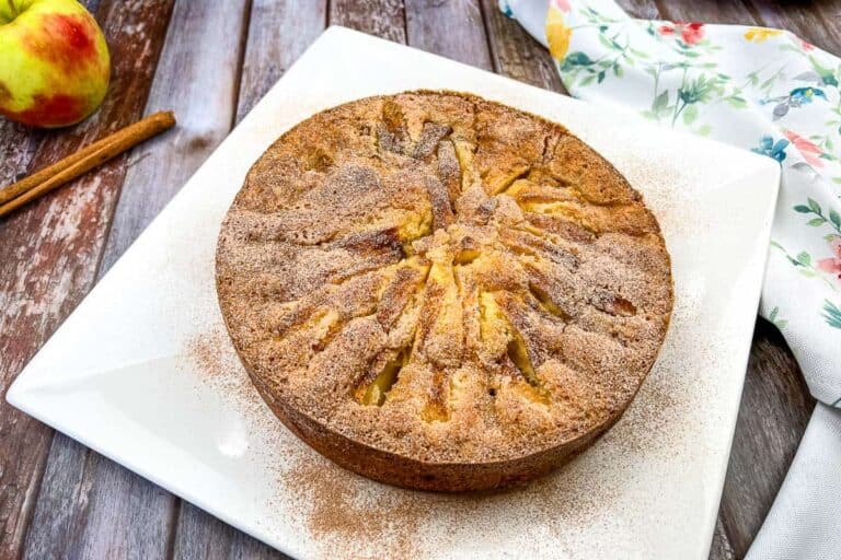 A round apple cake topped with cinnamon sugar sits on a white square plate, with a cinnamon stick, an apple, and a floral napkin nearby on a wooden table.