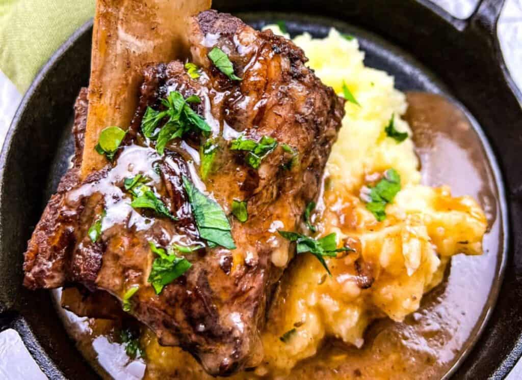 Beef short rib with gravy served over mashed potatoes, garnished with chopped parsley, in a black cast iron dish.