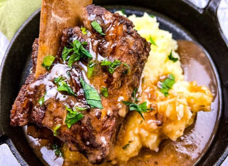 Beef short rib with gravy served over mashed potatoes, garnished with chopped parsley, in a black cast iron dish.