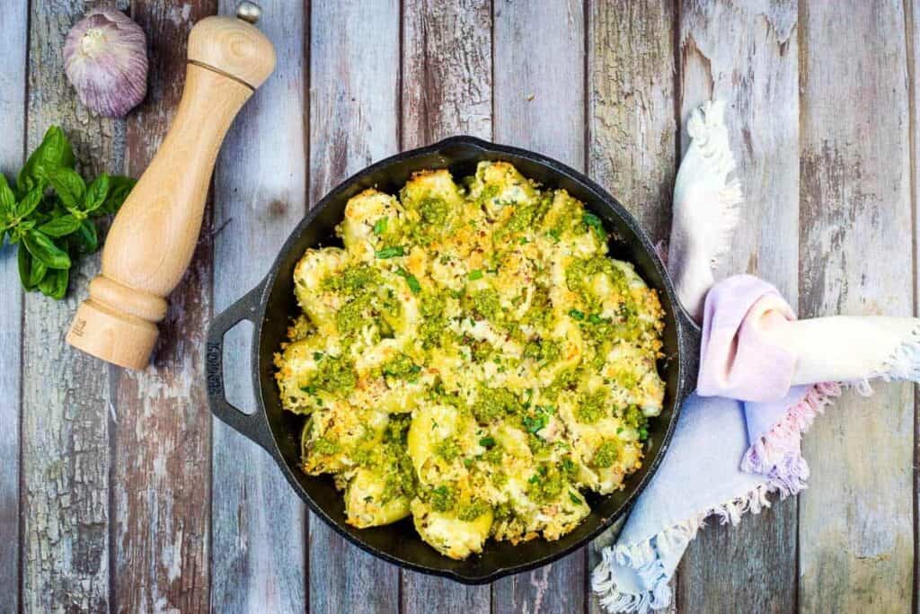 A cast iron skillet filled with baked pasta shells topped with green pesto and breadcrumbs sits on a rustic wooden table next to a napkin, basil leaves, a garlic bulb, and a pepper mill.