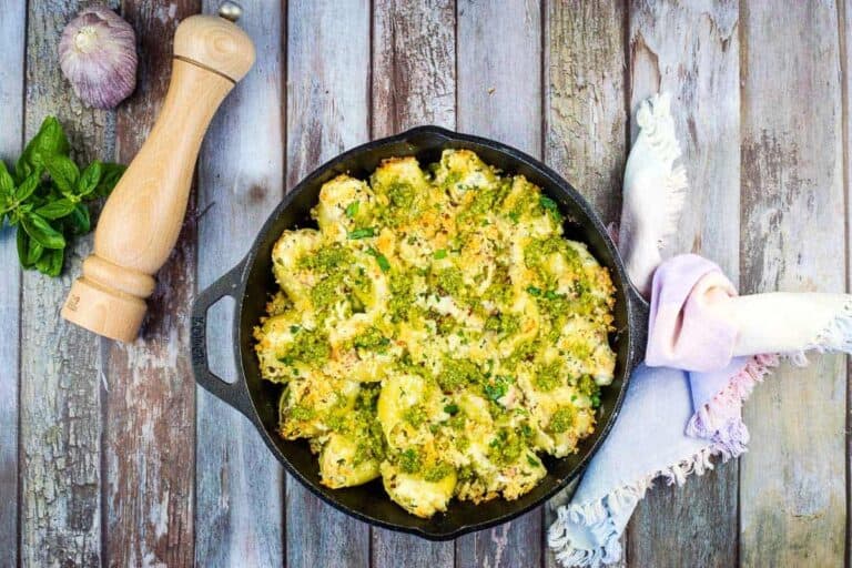 A cast iron skillet filled with baked pasta shells topped with green pesto and breadcrumbs sits on a rustic wooden table next to a napkin, basil leaves, a garlic bulb, and a pepper mill.
