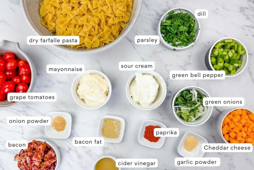 Ingredients for a pasta salad, including dry farfalle pasta, grape tomatoes, sour cream, mayonnaise, herbs, spices, bacon, green bell pepper, cheddar cheese, and seasonings displayed on a counter.