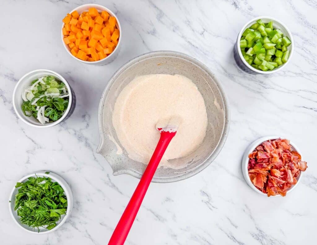 A mixing bowl of flour with a red spatula, surrounded by cups of chopped carrots, celery, green onions, herbs, and cooked bacon on a marble countertop.