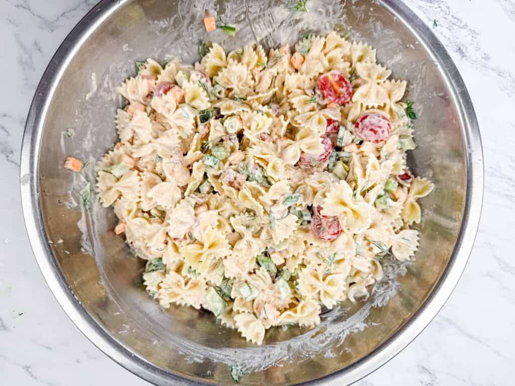 A stainless steel bowl filled with bowtie pasta salad mixed with vegetables and a creamy dressing, set on a white marble surface.