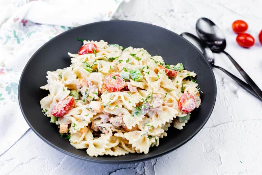 A black bowl filled with bowtie pasta, cherry tomatoes, chopped herbs, and a creamy sauce sits on a white surface next to two spoons and loose tomatoes.