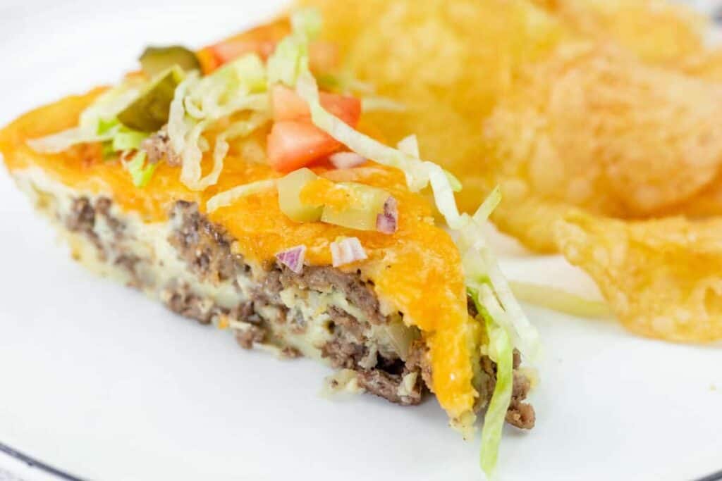 A slice of cheeseburger casserole topped with shredded lettuce, diced tomato, onion, and pickles, served with potato chips on a white plate.