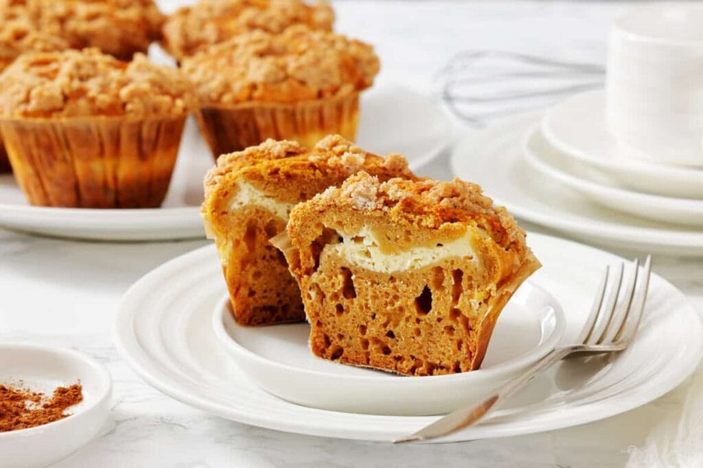 A plate with a pumpkin muffin cut in half, showing a cream cheese filling, next to a fork. Whole muffins and stacked dishes are in the background.