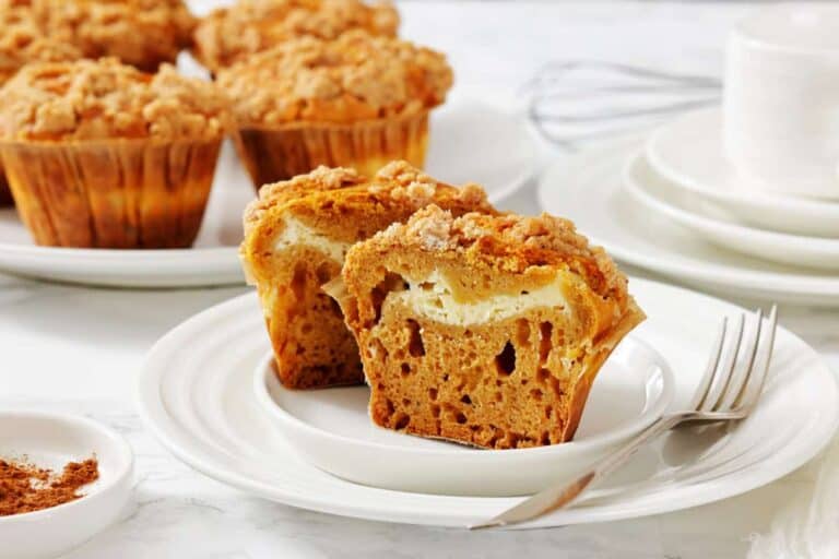 A plate with a pumpkin muffin cut in half, showing a cream cheese filling, next to a fork. Whole muffins and stacked dishes are in the background.