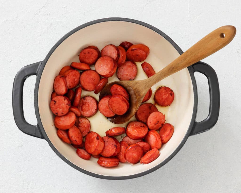 Sliced sausage being saut&eacute;ed in a white pot with a wooden spoon on a light surface.