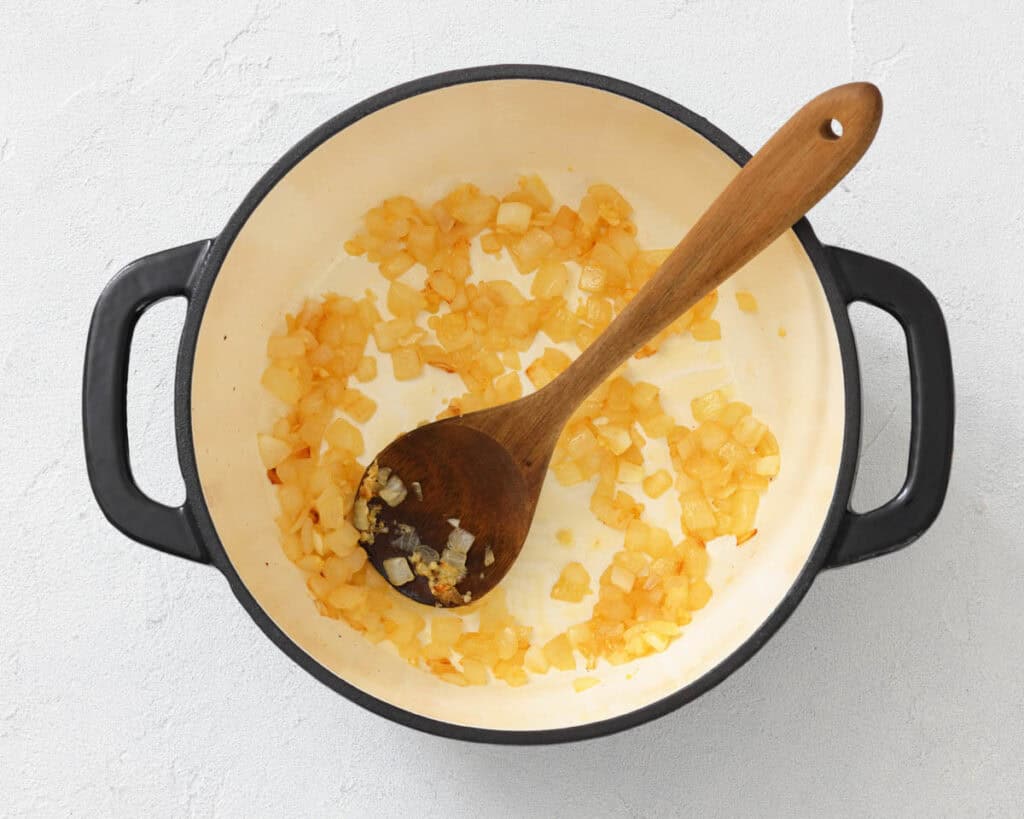 Diced onions saut&eacute;ing in a round, white enameled pot with black handles; a wooden spoon rests inside the pot.