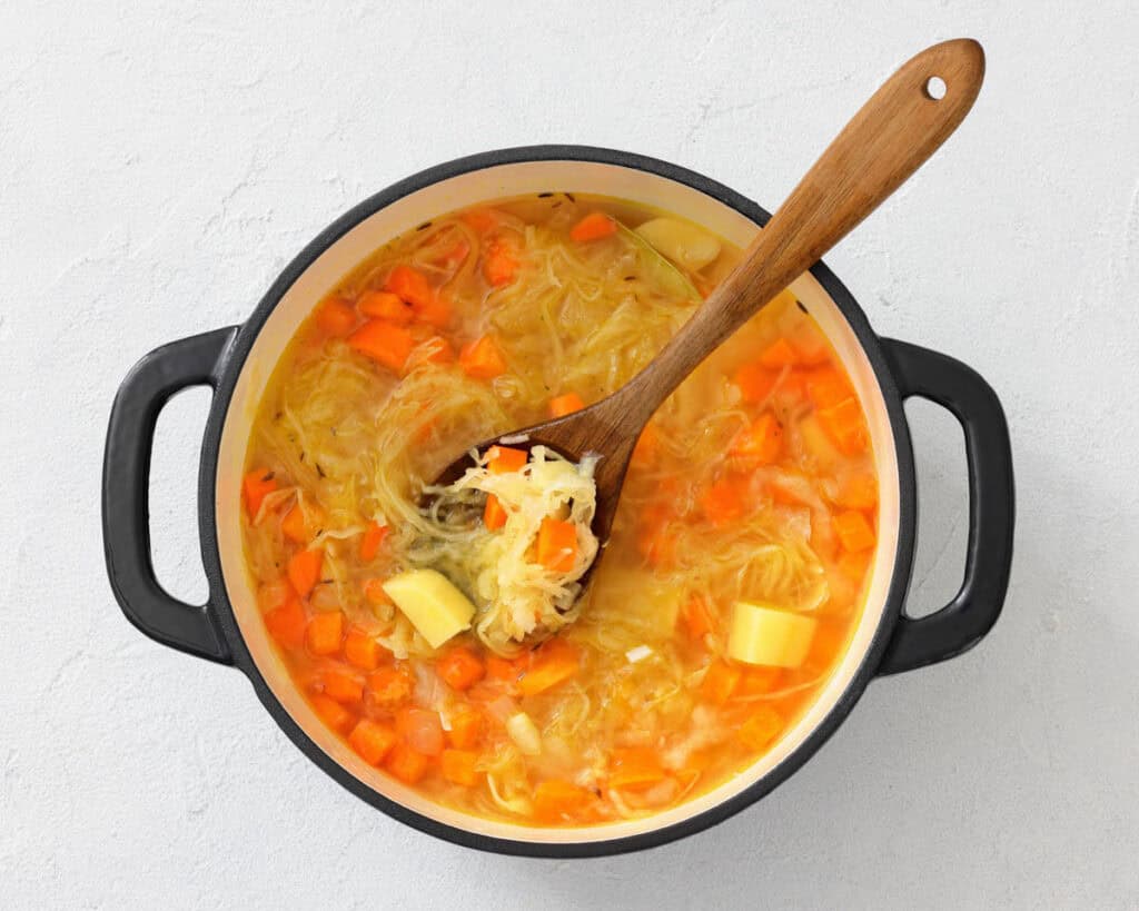 A pot of soup with chopped carrots, potatoes, and shredded cabbage, stirred with a wooden spoon on a light background.