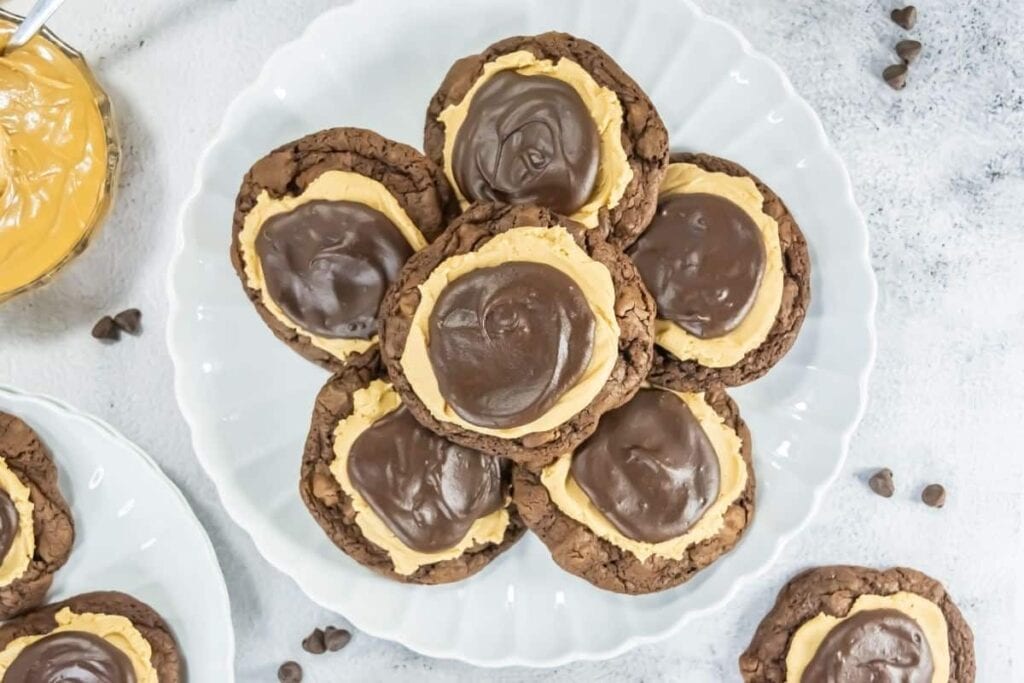A white plate with several chocolate cookies topped with a layer of peanut butter and a layer of chocolate frosting, surrounded by scattered chocolate chips.