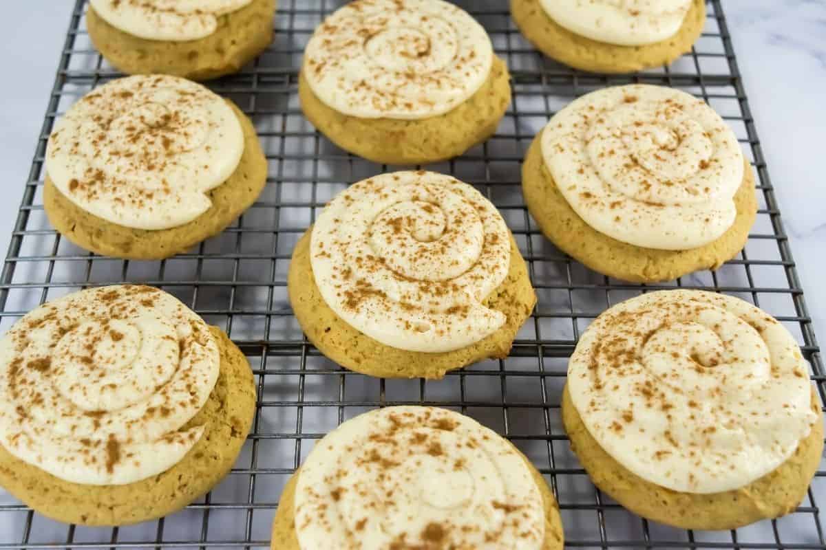 Copycat Crumbl Caramel Pumpkin Cookies with swirled white frosting and a sprinkle of cinnamon are cooling on a wire rack.