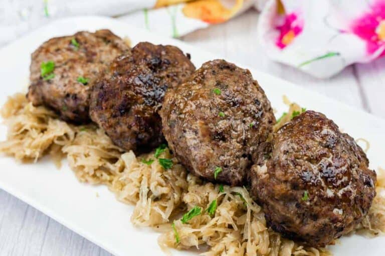 Four browned meat patties are arranged in a row on top of cooked shredded cabbage, garnished with a few green herbs, served on a rectangular white plate.
