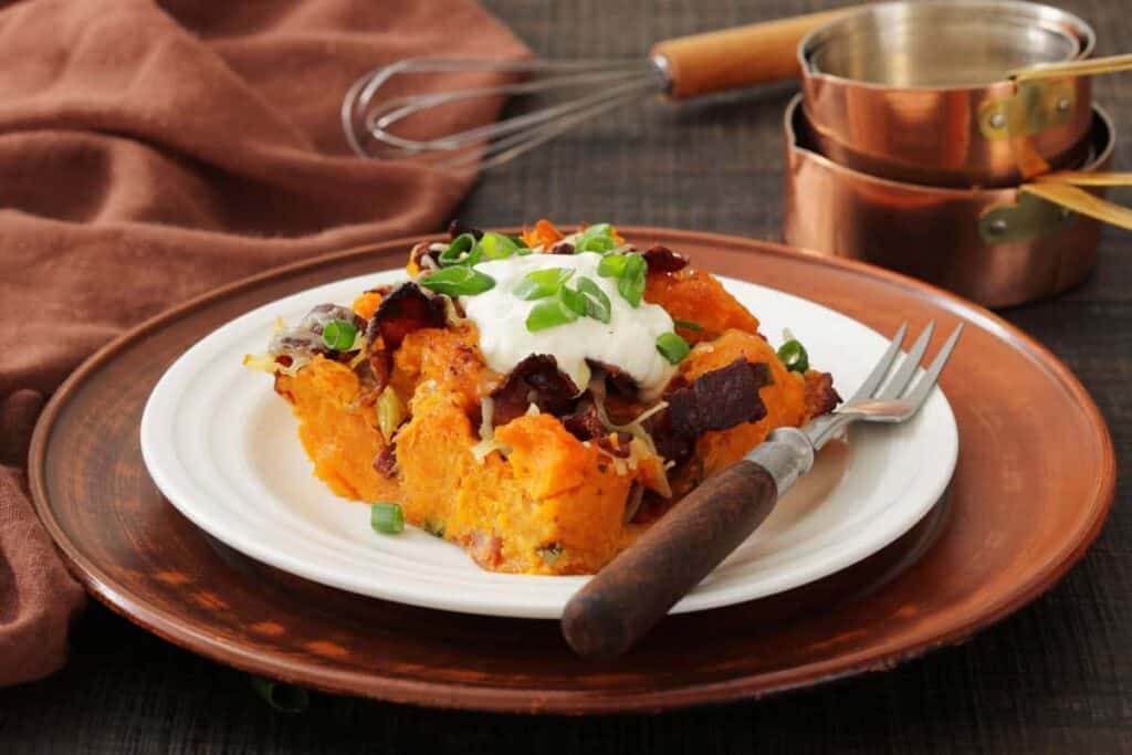 A plate of loaded sweet potato casserole topped with sour cream, chopped green onions, bacon bits, and cheese on a white dish with a fork. Copper measuring cups are in the background.