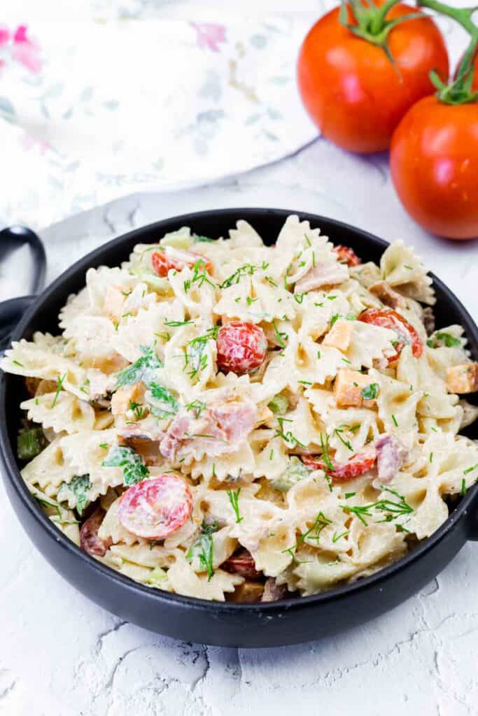 A black bowl filled with creamy pasta salad made with bowtie pasta, cherry tomatoes, herbs, and diced vegetables, with two whole tomatoes in the background.