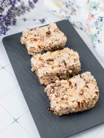 Three pieces of crumb-topped coffee cake with chopped nuts are arranged in a row on a rectangular black slate serving tray.