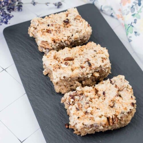 Three pieces of lazy daisy oatmeal cake with chopped nuts are arranged in a row on a rectangular black slate serving tray.