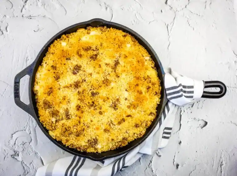 A cast-iron skillet filled with baked macaroni and cheese topped with golden breadcrumbs, resting on a gray textured surface with a striped kitchen towel.