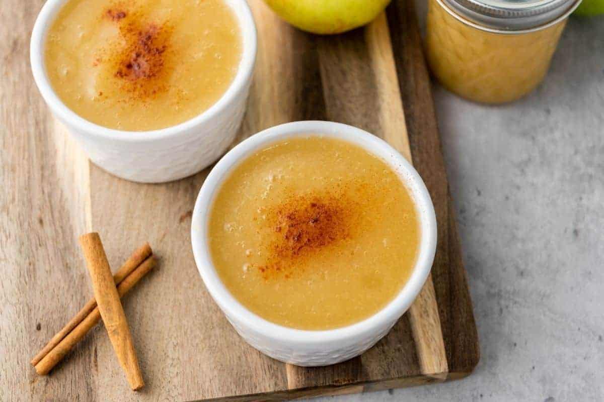 Two small white bowls of applesauce topped with cinnamon are placed on a wooden board next to cinnamon sticks, an apple, and a jar of applesauce.