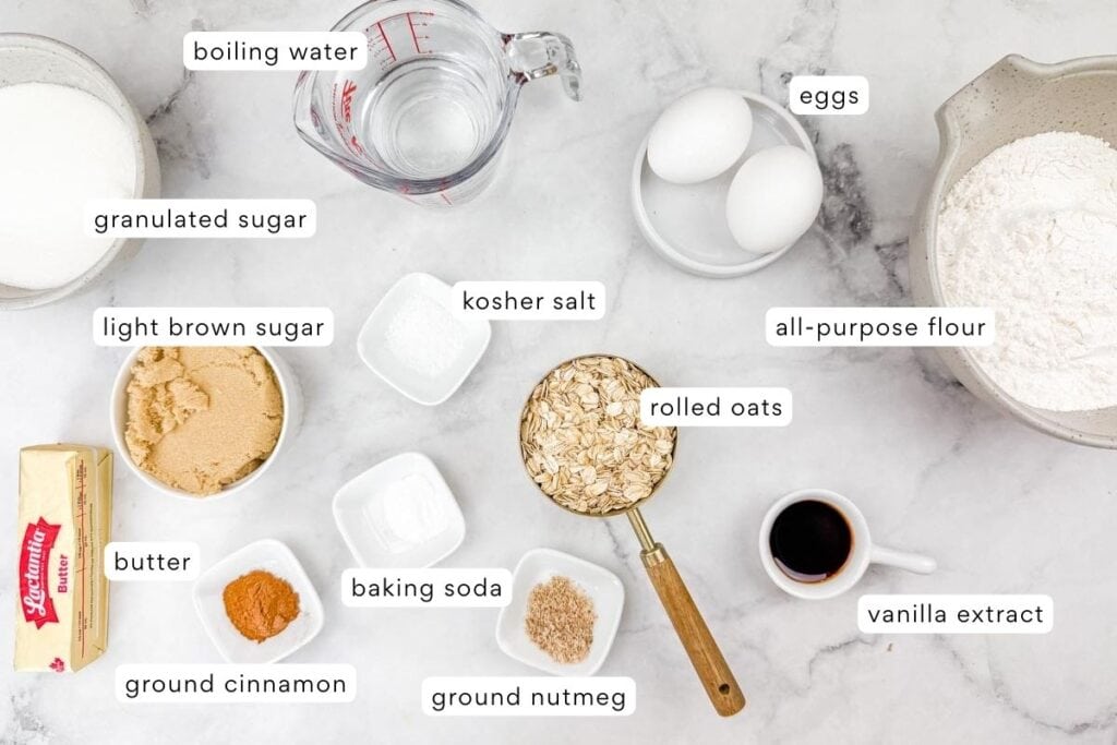 Baking ingredients including flour, eggs, rolled oats, sugars, butter, spices, vanilla extract, salt, baking soda, and a measuring cup of boiling water arranged on a marble surface.
