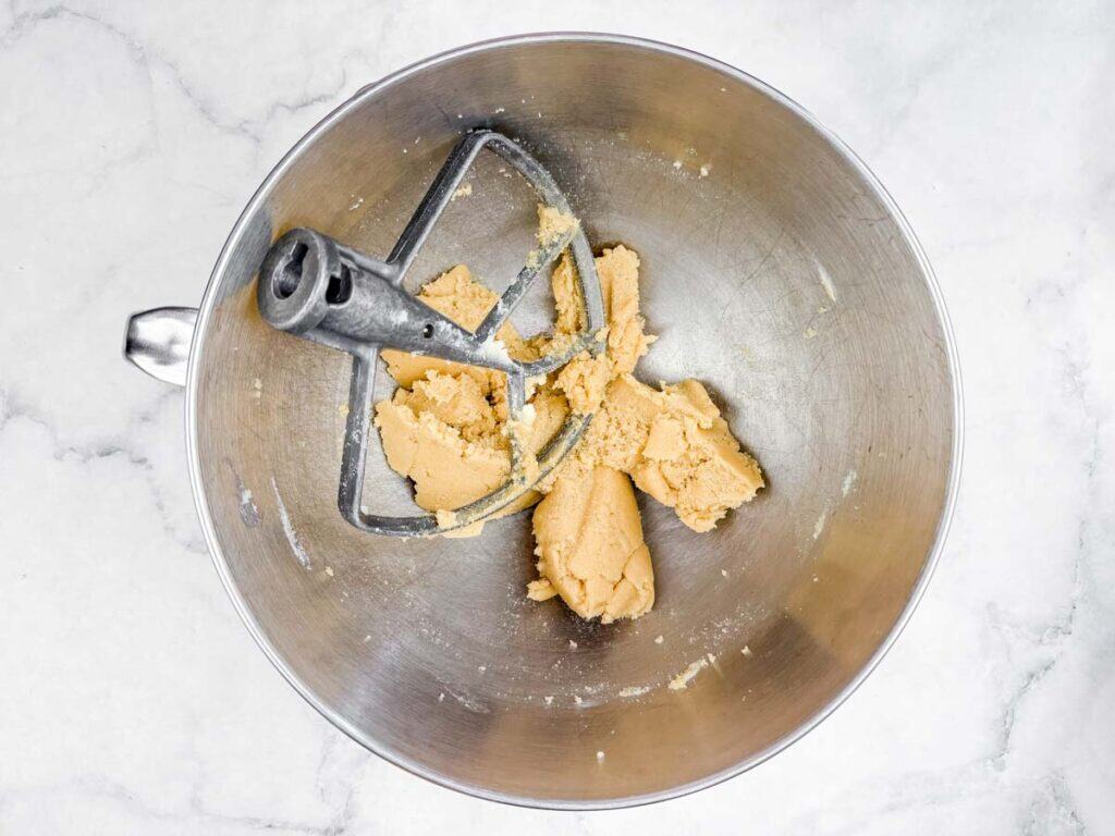 A metal mixing bowl with a paddle attachment contains partially mixed cookie dough on a marble surface.