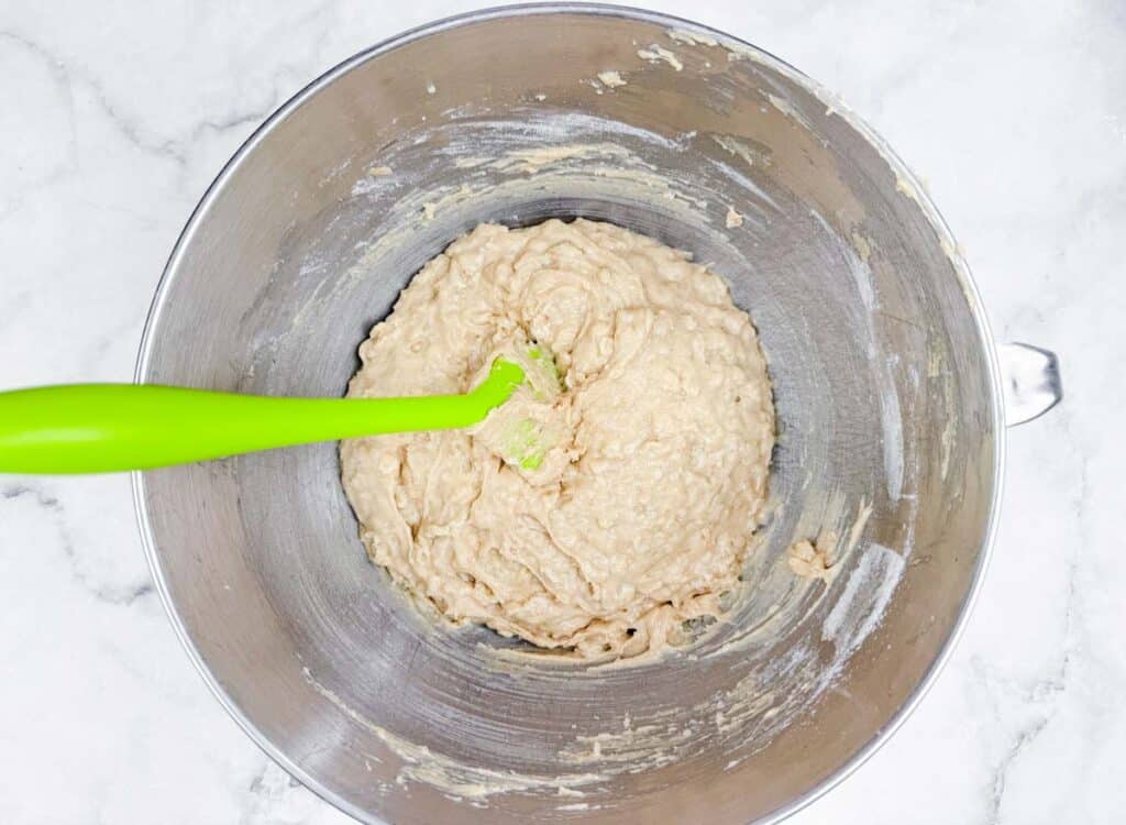A green spatula rests in a stainless steel mixing bowl filled with thick, beige batter on a white marble surface.
