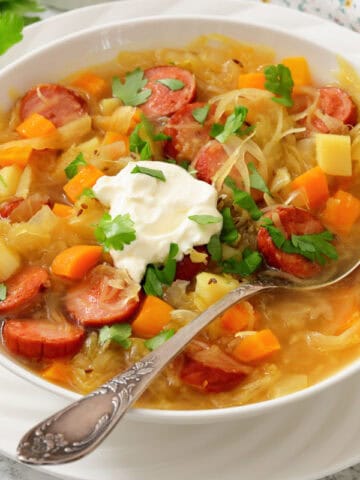 A bowl of sausage and vegetable soup topped with a dollop of sour cream and fresh herbs, with a spoon resting inside the bowl.