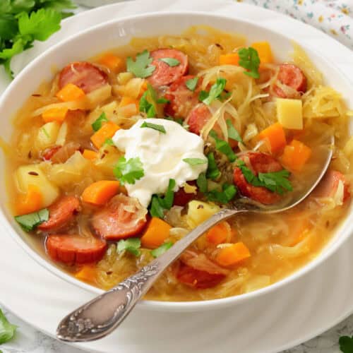 A bowl of sausage and vegetable soup topped with a dollop of sour cream and fresh herbs, with a spoon resting inside the bowl.