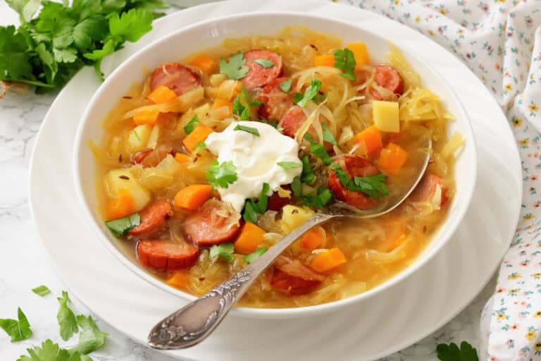 A bowl of kielbasa and vegetable soup topped with a dollop of sour cream and fresh herbs, with a spoon resting inside the bowl.