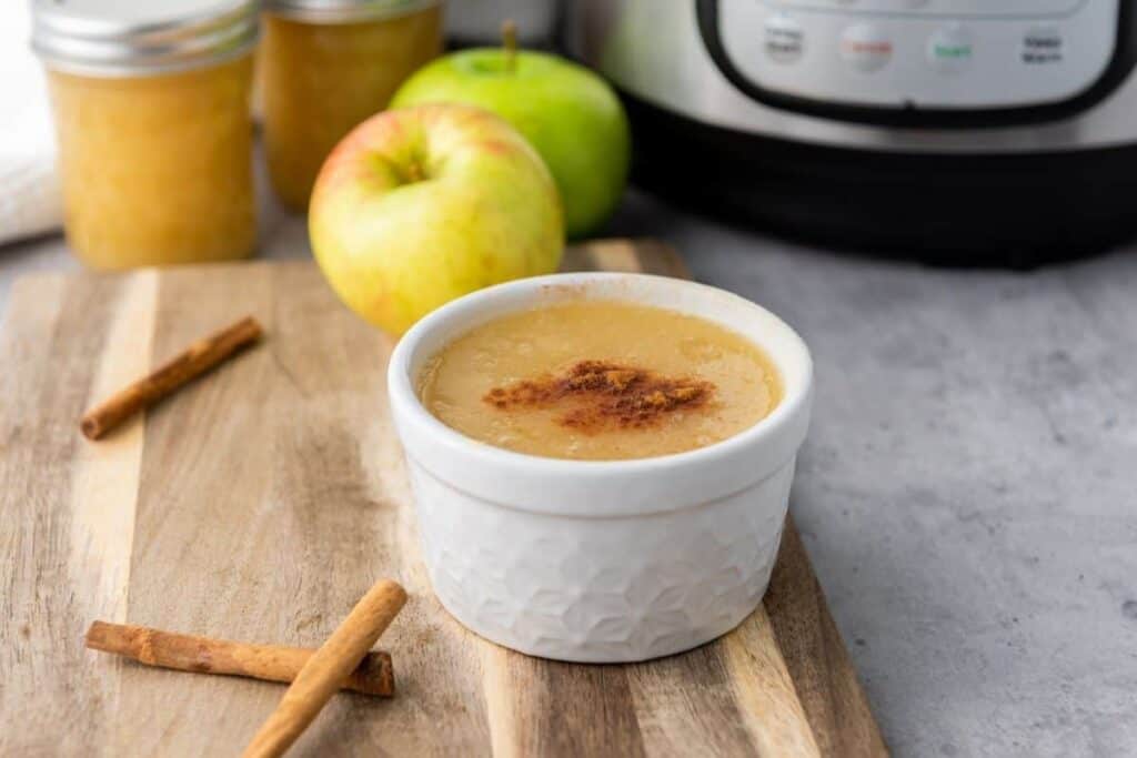 A white ramekin of applesauce topped with cinnamon sits on a wooden board with cinnamon sticks, apples, and jars in the background.