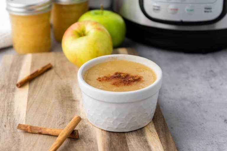 A white ramekin of applesauce topped with cinnamon sits on a wooden board with cinnamon sticks, apples, and jars in the background.