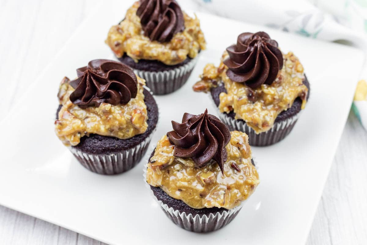 Four German Dark chocolate cupcakes topped with German chocolate coconut-pecan frosting and swirls of chocolate icing, arranged on a white square plate.