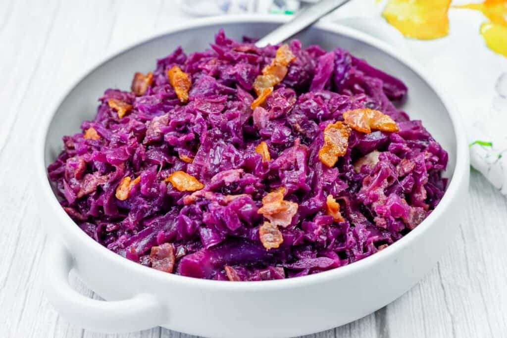 A white bowl filled with cooked red cabbage and pieces of crispy bacon, placed on a light wooden surface.