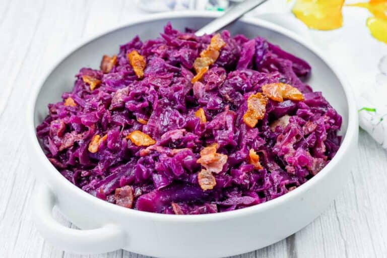 A white bowl filled with cooked red cabbage and pieces of crispy bacon, placed on a light wooden surface.