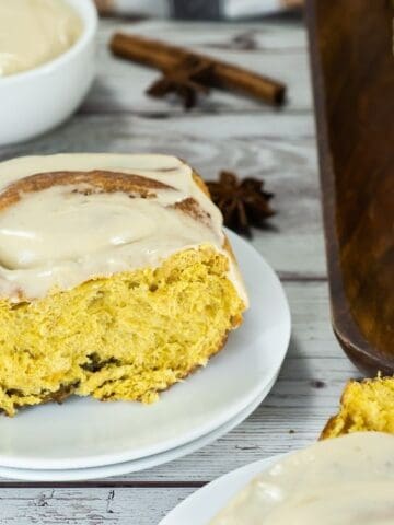A frosted cinnamon roll sits on a white plate, with more cinnamon rolls and a bowl of icing in the background on a wooden surface.