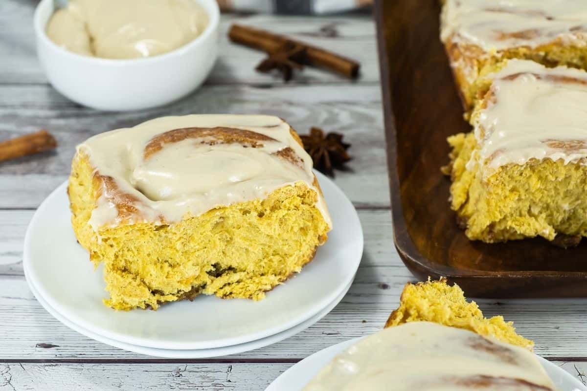 A frosted cinnamon roll sits on a white plate, with more cinnamon rolls and a bowl of icing in the background on a wooden surface.