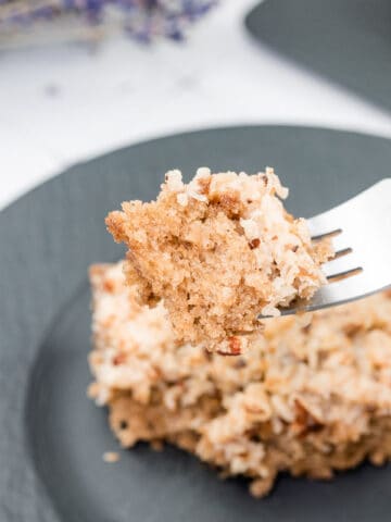 A fork holds a bite of crumbly, nut-topped cake above a black plate with a larger piece of the same cake.