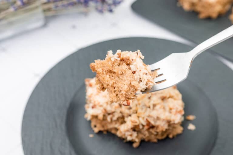 A fork holds a bite of crumbly, nut-topped cake above a black plate with a larger piece of the same cake.