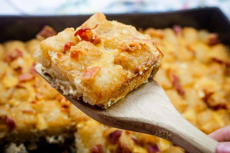 A wooden spatula holds a square piece of baked casserole topped with melted cheese and bits of bacon, with the rest of the casserole in the background.